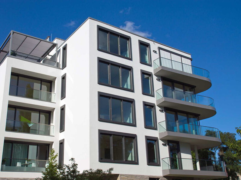 Modernes Mehrfamilienhaus mit großen Fenstern, Glasbalkonen und klarer Architektur vor blauem Himmel.